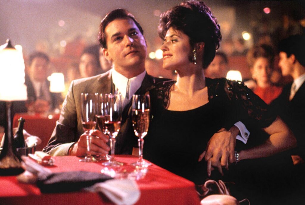 A film still from Goodfellas (1990) depicts actors Ray Liotta and Lorraine Bracco seated at a table in a nightclub.
