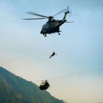 A helicopter lowers a rescue worker towards a gondola that is hanging precariously from a wire. The sun is setting and the gondola is positioned very high above the ground. There are mountains in the background.