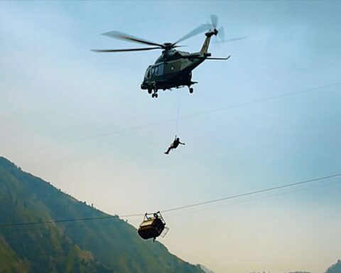 A helicopter lowers a rescue worker towards a gondola that is hanging precariously from a wire. The sun is setting and the gondola is positioned very high above the ground. There are mountains in the background.