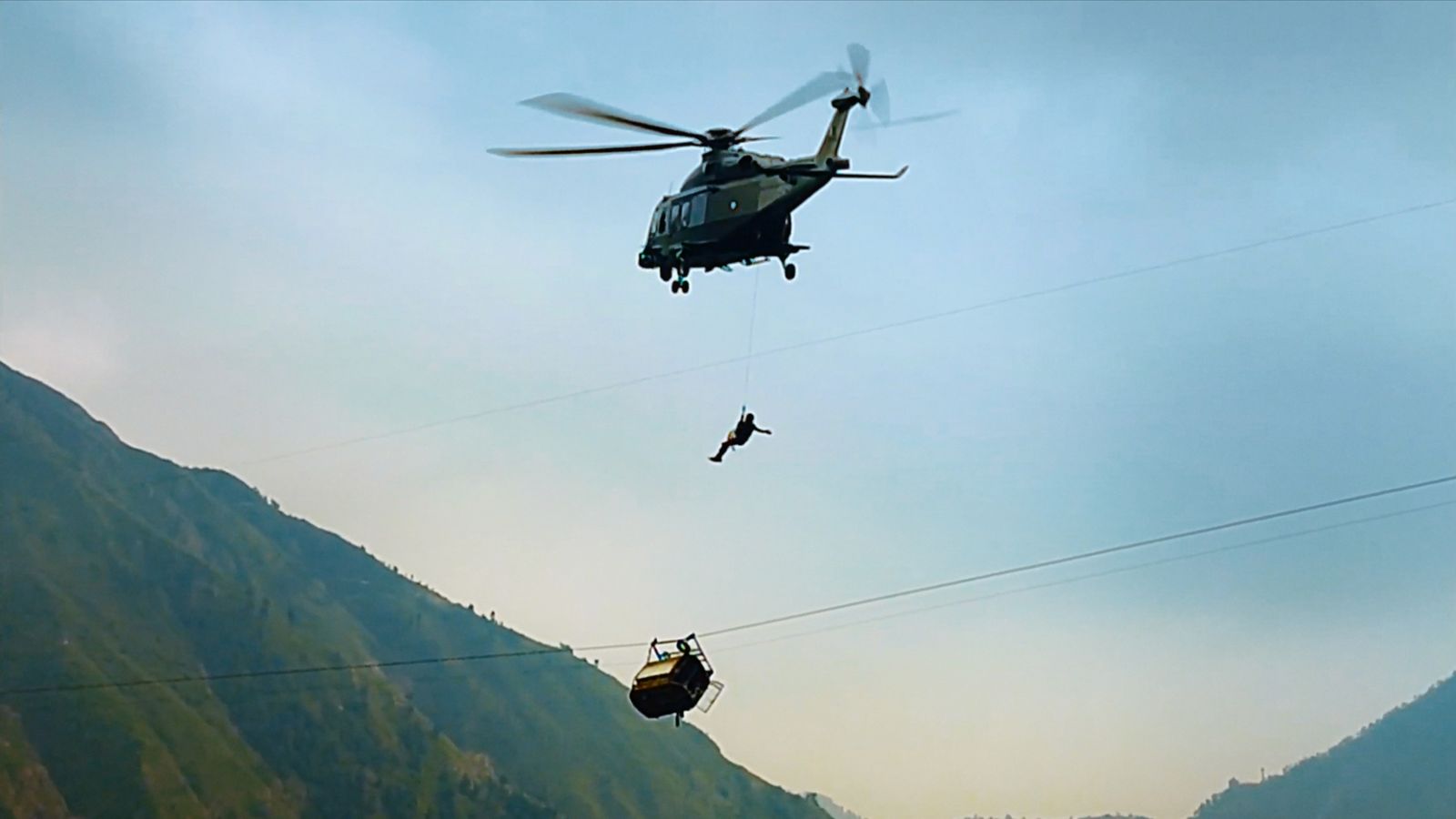 A helicopter lowers a rescue worker towards a gondola that is hanging precariously from a wire. The sun is setting and the gondola is positioned very high above the ground. There are mountains in the background.