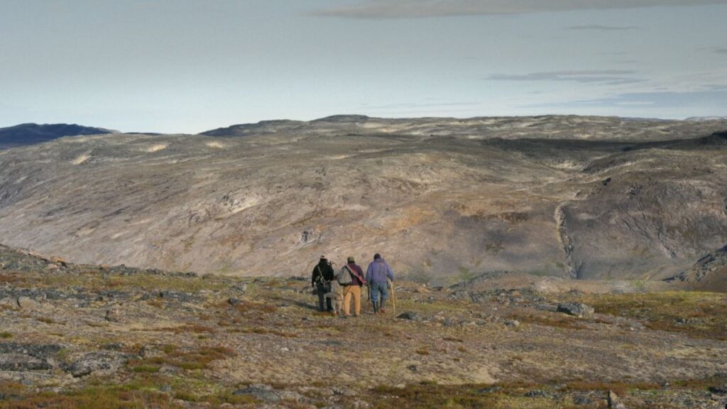 Three figures are seen from a distance as they stand in an Arctic landscape during the summer. The land is brown and green, and the sky is overcast.