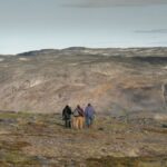 Three figures are seen from a distance as they stand in an Arctic landscape during the summer. The land is brown and green, and the sky is overcast.