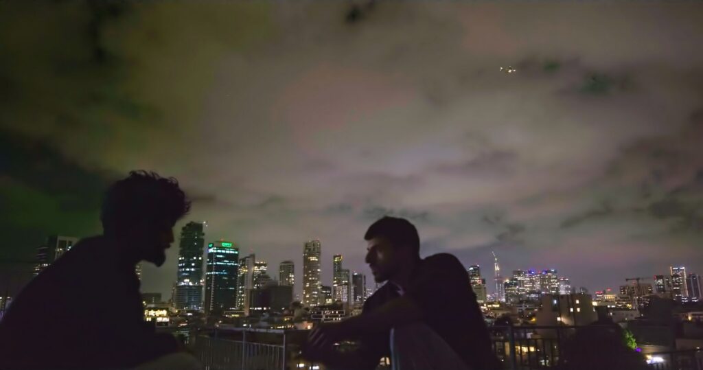 Two figures sit on a rooftop, facing each other, in conversation. It is a dark night and the figure on the left is completely obscured by shadow. On the right is an Israeli man.