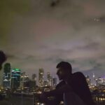 Two figures sit on a rooftop, facing each other, in conversation. It is a dark night and the figure on the left is completely obscured by shadow. On the right is an Israeli man.