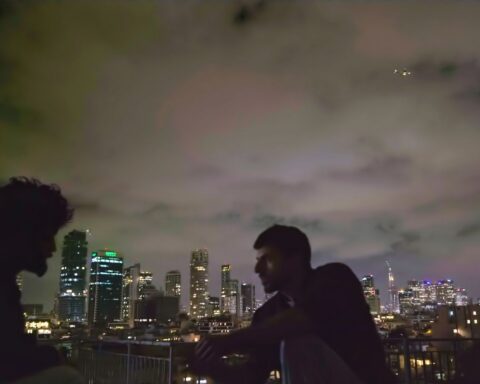 Two figures sit on a rooftop, facing each other, in conversation. It is a dark night and the figure on the left is completely obscured by shadow. On the right is an Israeli man.