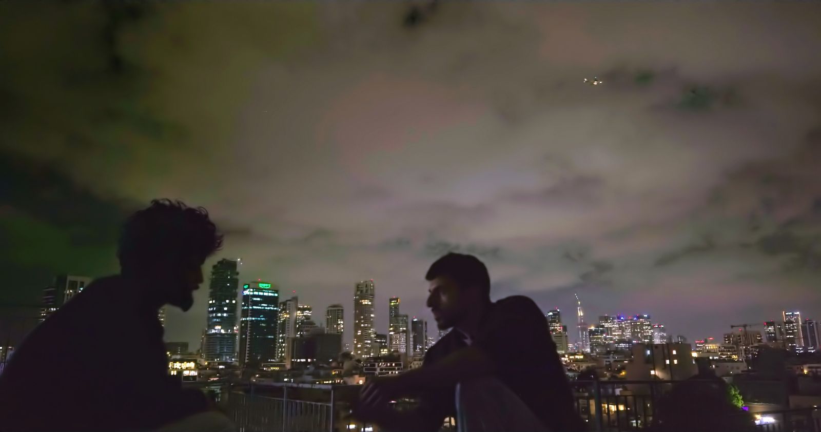Two figures sit on a rooftop, facing each other, in conversation. It is a dark night and the figure on the left is completely obscured by shadow. On the right is an Israeli man.