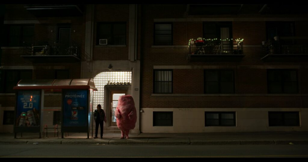 A woman stands on a darkened street at a bus stop. A large cotton candy-like pink figure stands beside her.