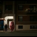A woman stands on a darkened street at a bus stop. A large cotton candy-like pink figure stands beside her.
