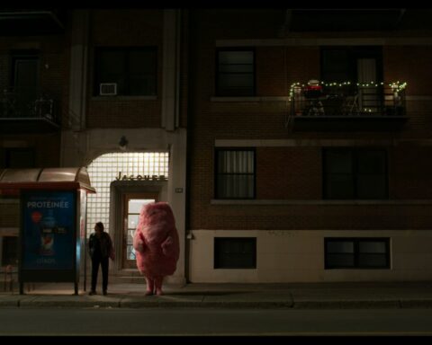 A woman stands on a darkened street at a bus stop. A large cotton candy-like pink figure stands beside her.