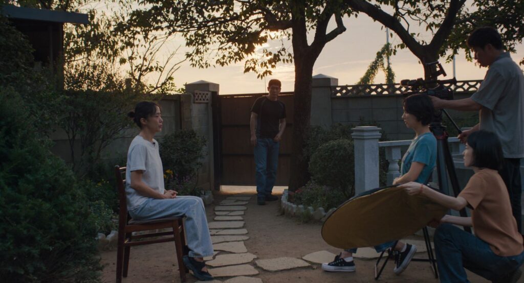 Two South Korean women sit and conduct an interview. The woman on the left is wearing a white shirt and faces the camera, and the woman on the right wears a green shirt and sits beside the camera while asking questions. There is a film crew behind her, and a man stands between them in the background observing the interview. They are in a backyard and the sun is setting.