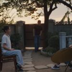 Two South Korean women sit and conduct an interview. The woman on the left is wearing a white shirt and faces the camera, and the woman on the right wears a green shirt and sits beside the camera while asking questions. There is a film crew behind her, and a man stands between them in the background observing the interview. They are in a backyard and the sun is setting.