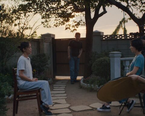Two South Korean women sit and conduct an interview. The woman on the left is wearing a white shirt and faces the camera, and the woman on the right wears a green shirt and sits beside the camera while asking questions. There is a film crew behind her, and a man stands between them in the background observing the interview. They are in a backyard and the sun is setting.