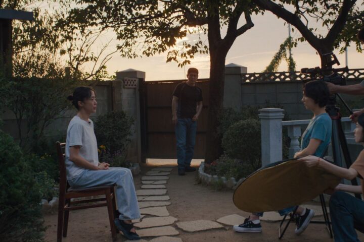 Two South Korean women sit and conduct an interview. The woman on the left is wearing a white shirt and faces the camera, and the woman on the right wears a green shirt and sits beside the camera while asking questions. There is a film crew behind her, and a man stands between them in the background observing the interview. They are in a backyard and the sun is setting.
