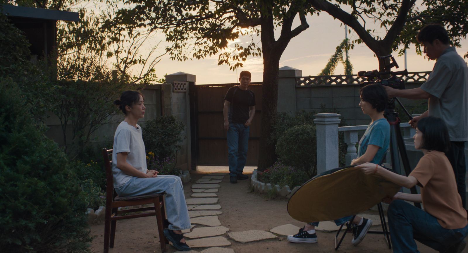 Two South Korean women sit and conduct an interview. The woman on the left is wearing a white shirt and faces the camera, and the woman on the right wears a green shirt and sits beside the camera while asking questions. There is a film crew behind her, and a man stands between them in the background observing the interview. They are in a backyard and the sun is setting.