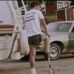 An archival image shows Terry Fox from behind. He is wearing a white t-shirt and black jogging shorts. He is holding a prosthetic leg in his hands. There is a white camper van beside him.