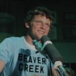An archival image shows Terry Fox in close-up as he stands in front of a microphone. He has a smile on his face and is wearing a blue shirt with black letters that read "Beaver Creek".