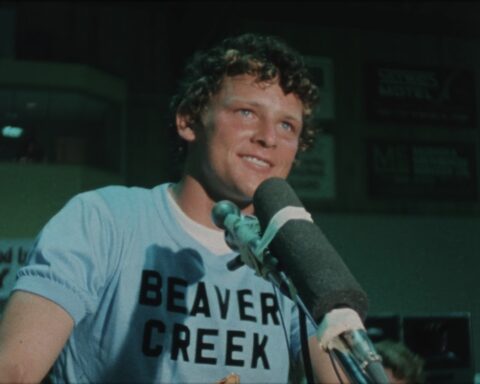 An archival image shows Terry Fox in close-up as he stands in front of a microphone. He has a smile on his face and is wearing a blue shirt with black letters that read "Beaver Creek".