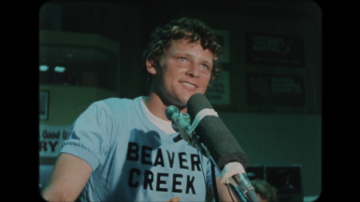 An archival image shows Terry Fox in close-up as he stands in front of a microphone. He has a smile on his face and is wearing a blue shirt with black letters that read "Beaver Creek".