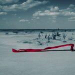 A long shot displays a person walking through a desert and holding a long billowing piece of red fabric that flows in the wind behind them.