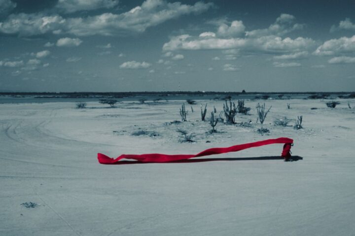 A long shot displays a person walking through a desert and holding a long billowing piece of red fabric that flows in the wind behind them.