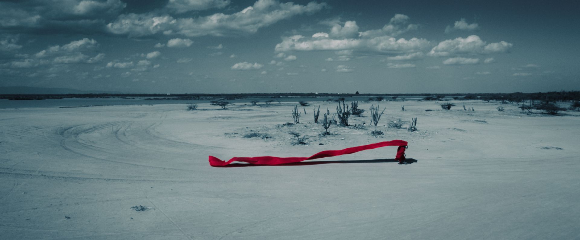 A long shot displays a person walking through a desert and holding a long billowing piece of red fabric that flows in the wind behind them.