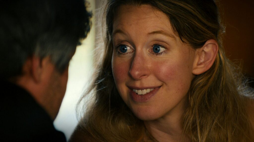 A close-up image of Elizabeth Holmes shows a wide-eyed expression and a mysterious smile. She is a blonde white woman with blue eyes, pictured in close-up, speaking to someone who is positioned in front of the camera.