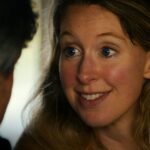 A close-up image of Elizabeth Holmes shows a wide-eyed expression and a mysterious smile. She is a blonde white woman with blue eyes, pictured in close-up, speaking to someone who is positioned in front of the camera.