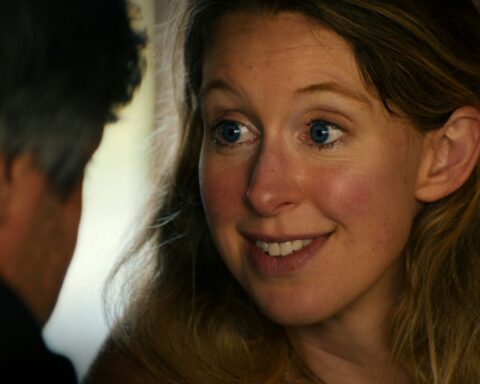 A close-up image of Elizabeth Holmes shows a wide-eyed expression and a mysterious smile. She is a blonde white woman with blue eyes, pictured in close-up, speaking to someone who is positioned in front of the camera.